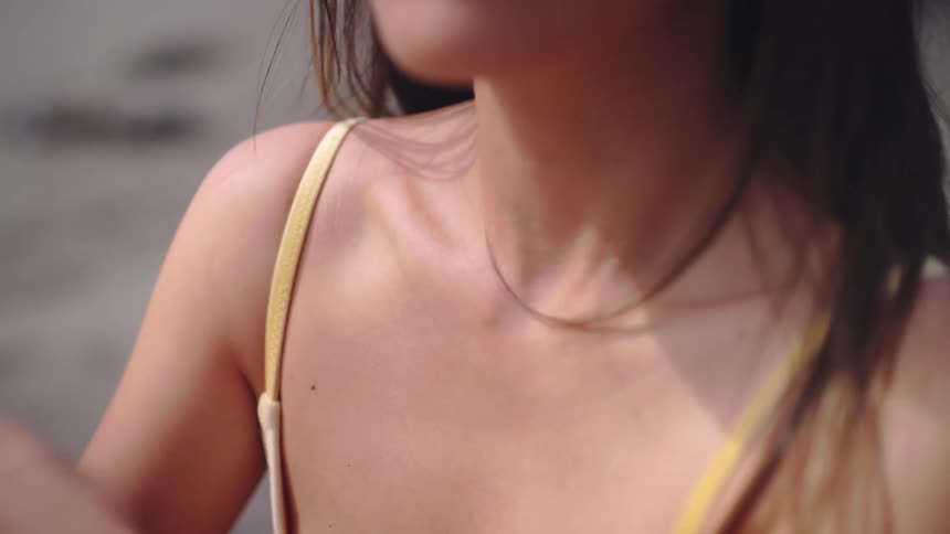 A close view of a womans shoulder and neck shows her in a yellow strapped top touching her collarbone and shoulder with her hand in soft outdoor sunlight