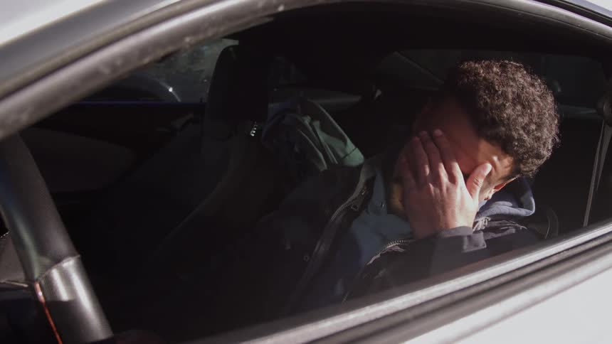 A tired looking man in a dark jacket wakes up in the drivers seat of a parked car and then rubs his face with his hands shielding his eyes from the light