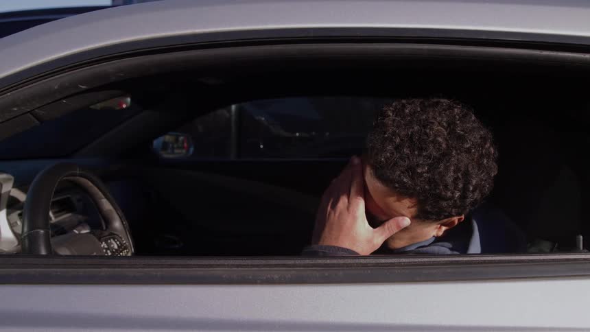 A man sits in the drivers seat of a silver car parked on a city street first resting his chin on his fist then rubbing his forehead and covering his face with his hand in an expression of fatigue