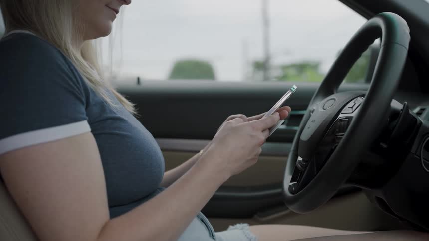 A young woman sits behind the steering wheel of a car texting on her mobile phone at midday