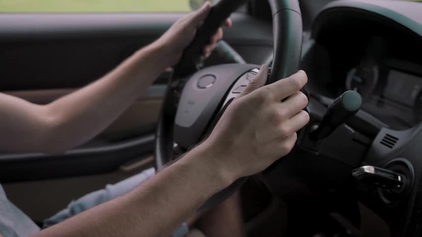 A close view inside a car shows the hands of a man gripping the steering wheel as he drives in the daylight