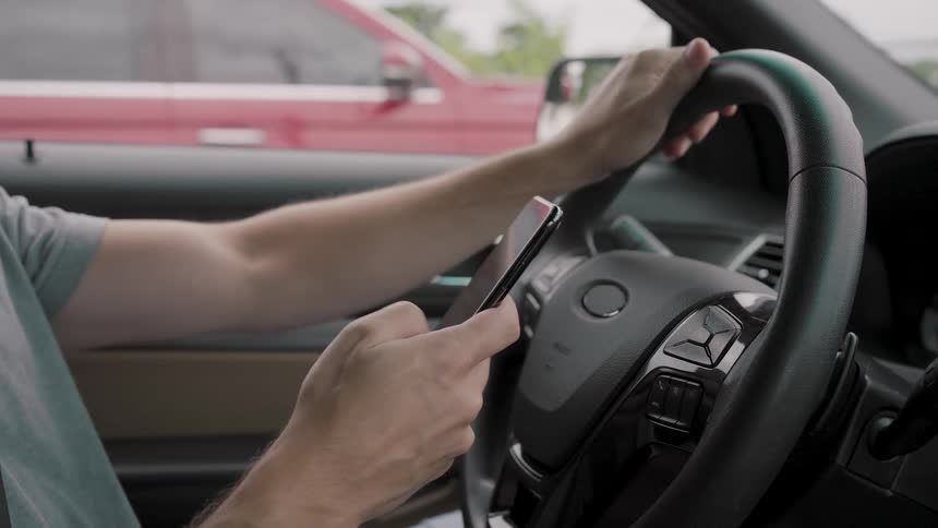 A closeup shot from inside a car shows a person using a smartphone with one hand while the other hand rests on the steering wheel the vehicle appears to be stopped with a red truck visible through the windshield in the background