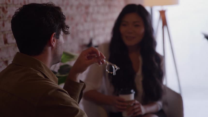The video shows two people having a conversation indoors in a modern comfortable settinga man in the foreground seen from behind the shoulder is holding a pair of glasses as he looks toward a smiling woman who is speaking to him and holding
