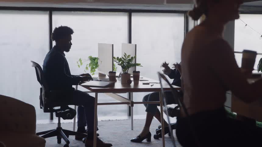 A group of office workers are silhouetted against bright large windows in a modern office as a man works on his computer and a woman sits opposite him talking and gesturing