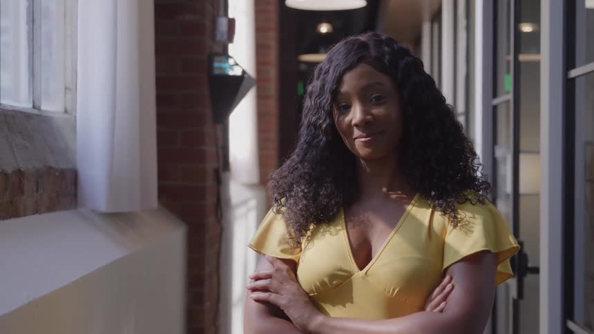 A woman with long dark curly hair stands in a brightly lit hallway smiling confidently with her arms crossed she is wearing a yellow top and is positioned next to a large window suggesting an office or school building environment
