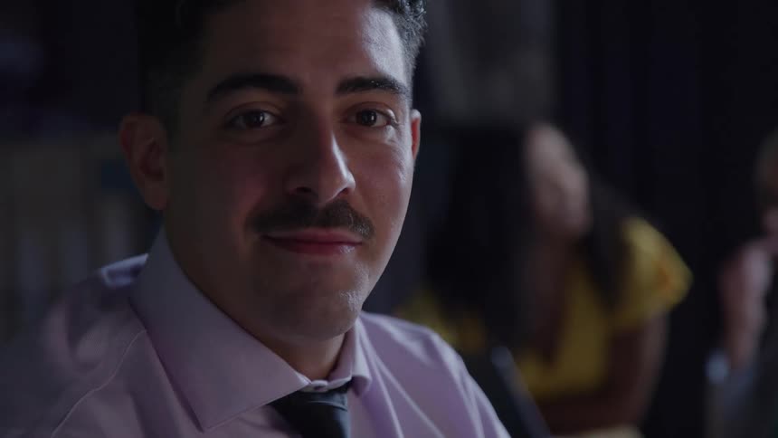 Man with a mustache wearing a white shirt and a black tie sitting down and smiling with two women talking on the background indoors
