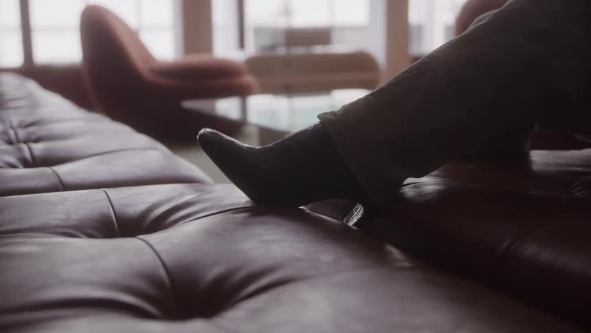 Person seated with crossed leg on brown leather couch in naturally lit room