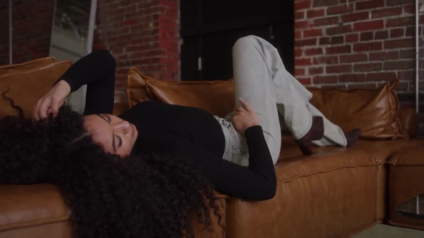 A woman with long curly dark hair wearing a black long sleeve shirt and light gray pants with boots lies on her side on a brown leather couch in a cozy industrial living space