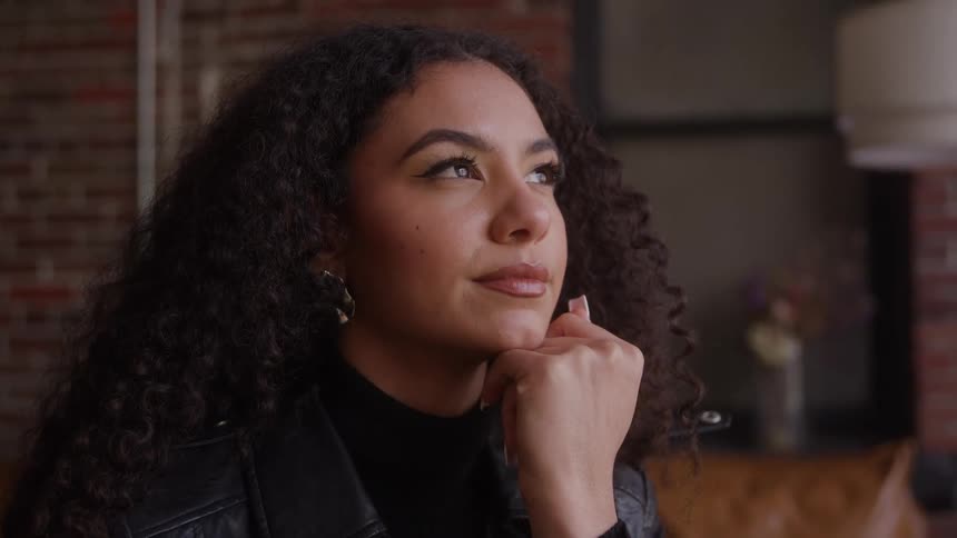 A young black woman sits indoors resting one hand under her chin with long curly black hair and a smile on her face wearing a black leather jacket