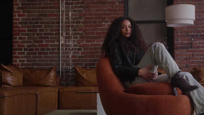 Woman with curly hair sits in modern chair holding phone in indoor space with brick wall and leather sofa in the background
