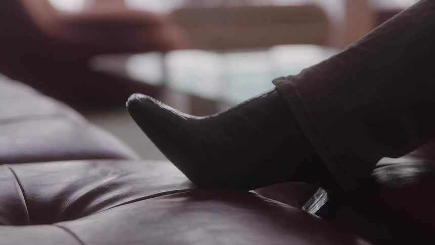 A booted high heel foot rests on a leather couch in a softly lit living room