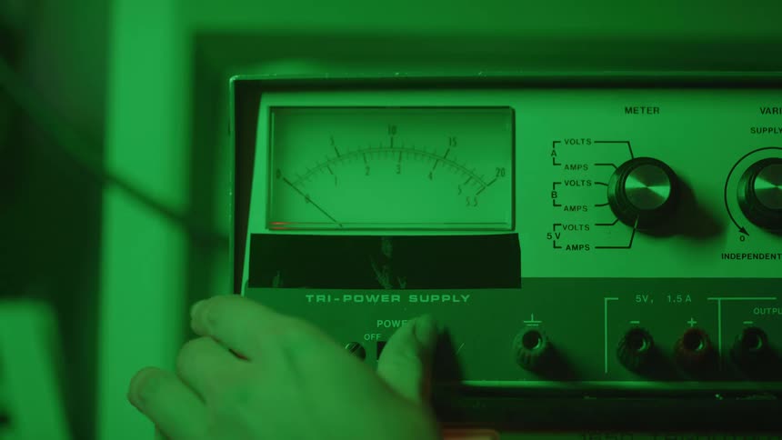 A hand flips the power switch of an old analog tri power supply machine with a glowing green meter and an illuminated red power light set in a dark room with a green color tint