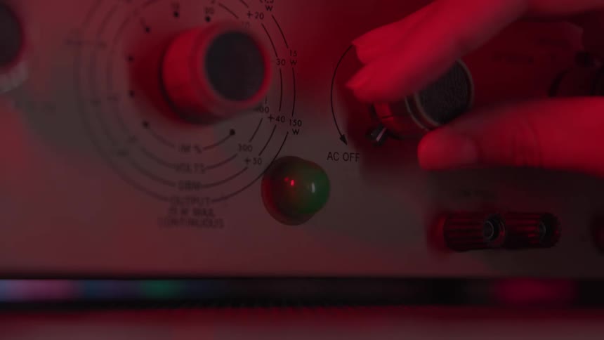 A hand turns a knob on a vintage control panel under red light that turns green