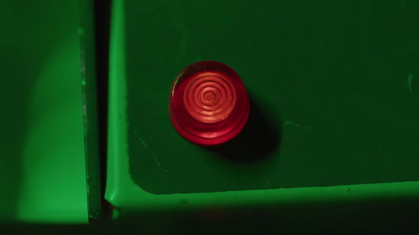 A flashing small red circular button with concentric rings sits on a dark green metal surface