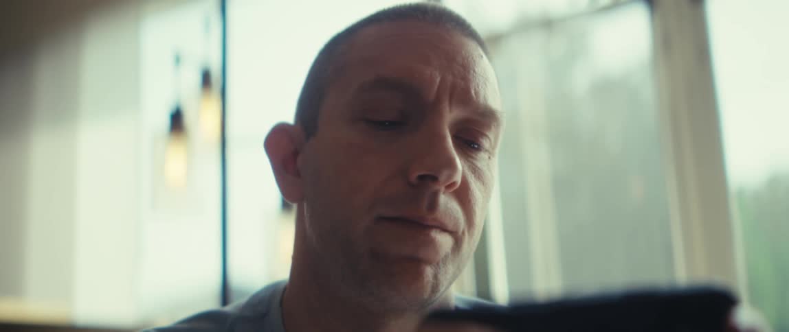 The video is an extreme closeup of a mans face in an indoor setting with bright natural lighting coming from windows in the background he has a serious or concentrated expression as he holds a dark object possibly a phone or remote control i