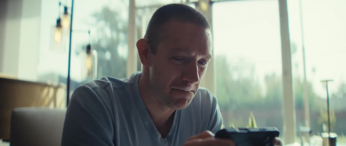 A man with short hair sits indoors and looks down with concentration at a black smartphone he holds horizontally and bright natural light comes through a large window behind him