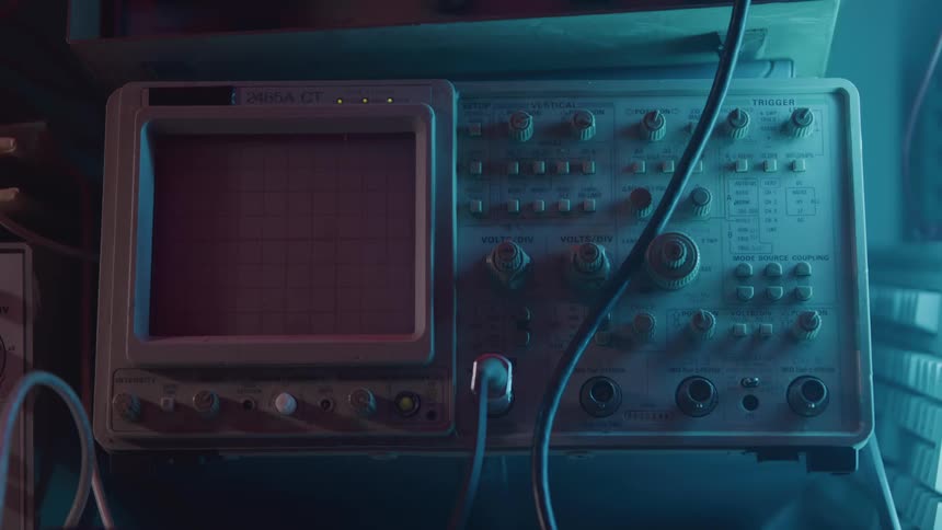 Hand adjusts an oscilloscope displaying green signals on a red screen under bluish light