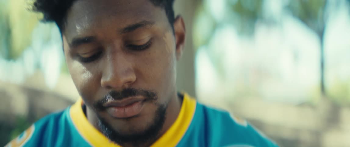 A closeup shot of a man wearing a blue and yellow sports jersey shows him looking thoughtful and slightly downcast as he briefly closes his eyes the camera moves down to his chest where he then raises his phone into view