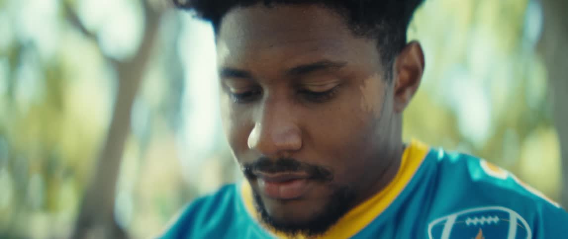 A man wearing a blue and yellow jersey holds a phone in his hands he looks down at the screen with a subtle smile and then a serious expression and he continues to interact with the device while sitting on a bench outdoors