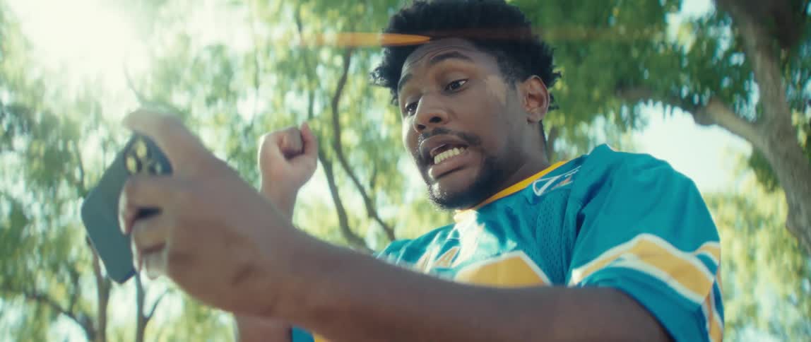 A man wearing a blue and yellow sports jersey is outdoors in a park intently holding his phone horizontally as he plays a mobile game he reacts with an openmouthed expression of surprise or excitement then laughs and pumps his fist in a clear celebrat
