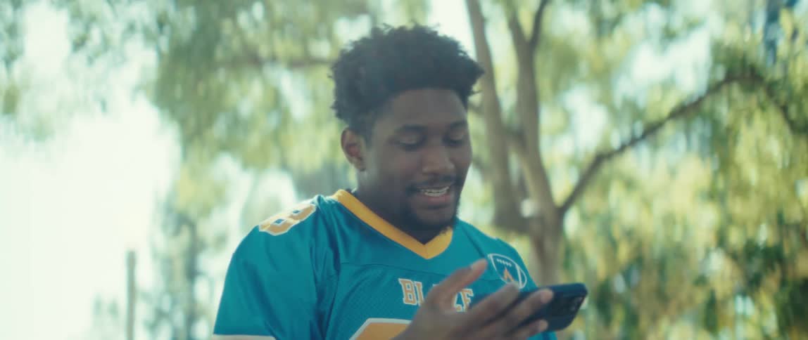 A young man wearing a jersey walks through a public park at midday watching content on his mobile device with excitement