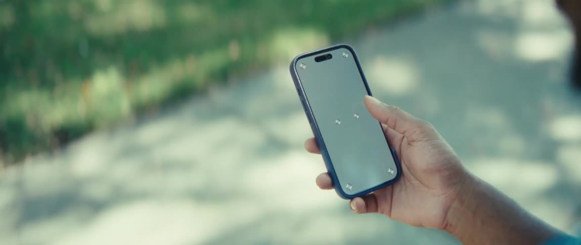 A young man operating his iphone standing under a tree in a public park on a sunny day