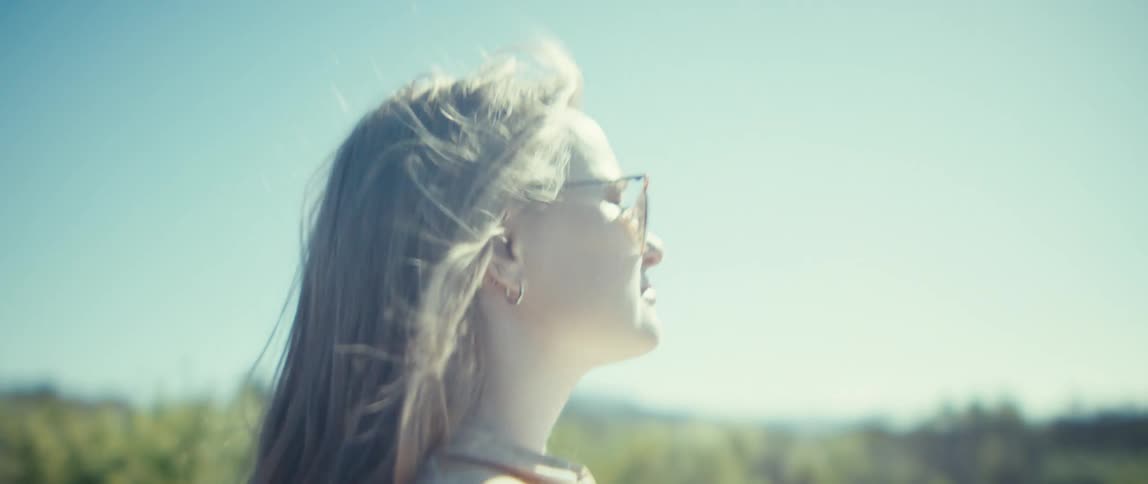 A bright sunlit close view shows a woman wearing sunglasses and a patterned shirt walking in an open dry field