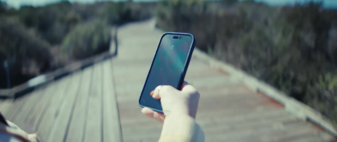 A person stands on an outdoor wooden boardwalk she holds a darkcased smartphone upright in one hand she lightly touches the screen with her thumb and the blurred boardwalk recedes into the background