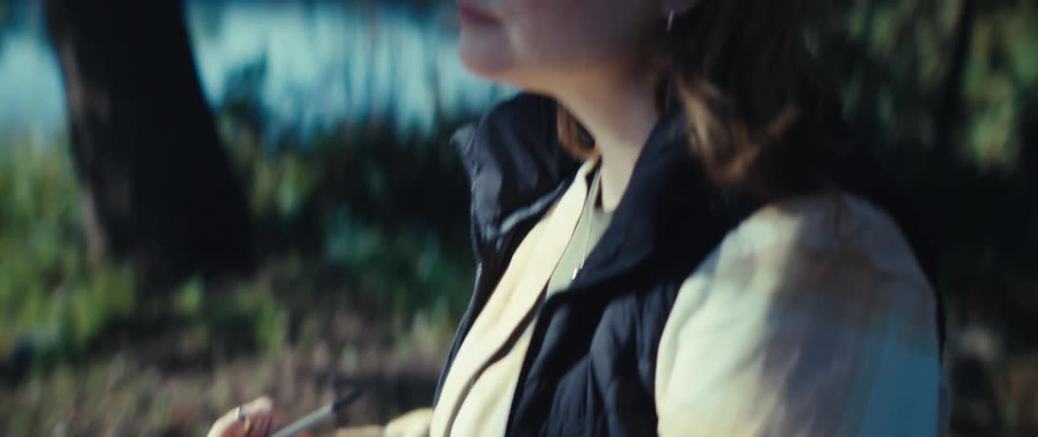 A woman is sitting outside by a lake and sketching