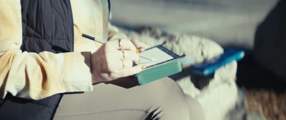 The video begins with a medium shot of a woman outdoors wearing glasses and a dark vest over a lightcolored topit then transitions to a closeup focusing on her hands as she uses a pen to write or sketch in a small green notebook resting on