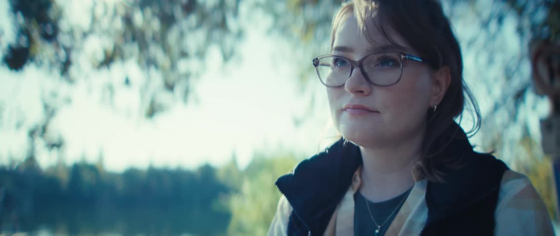 A young woman wearing glasses writes with a pen under a tree in the wild at midday