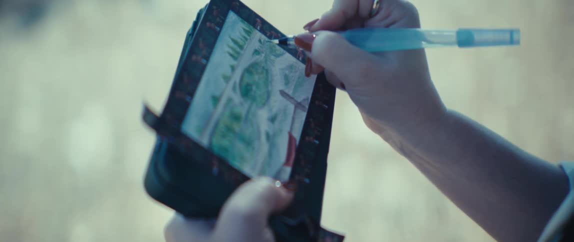 Hands hold a piece of paper framed with a drawing on it a tool is used to add lines to the artwork and the background is blurry under sunlight
