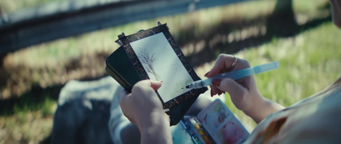 A woman sitting on an outdoor bench painting on a small piece of paper