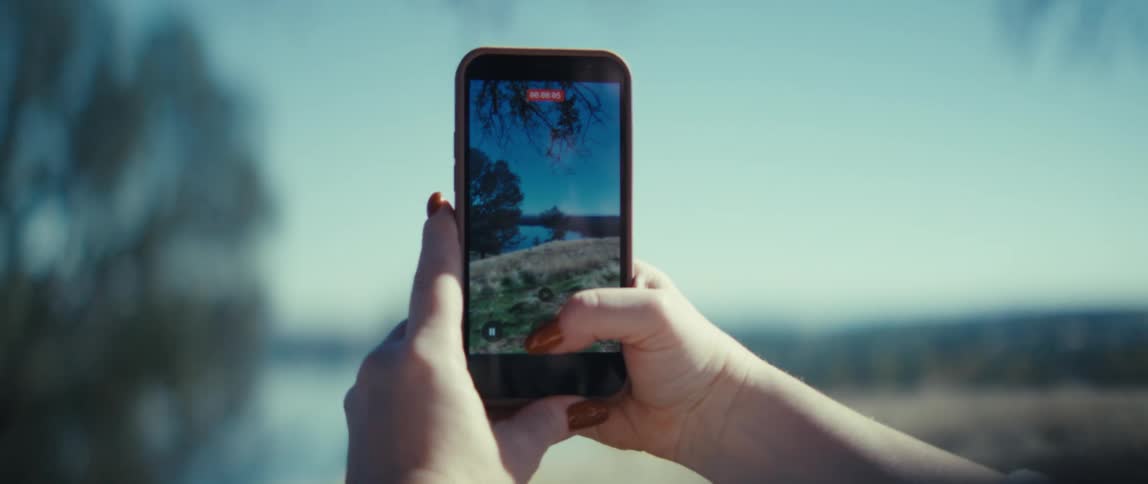 The video is a closeup of a persons hands holding a smartphone vertically to record a video of an outdoor landscapethe screen shows a scene with bare tree branches against a blue sky above a dry grassy and hazy field