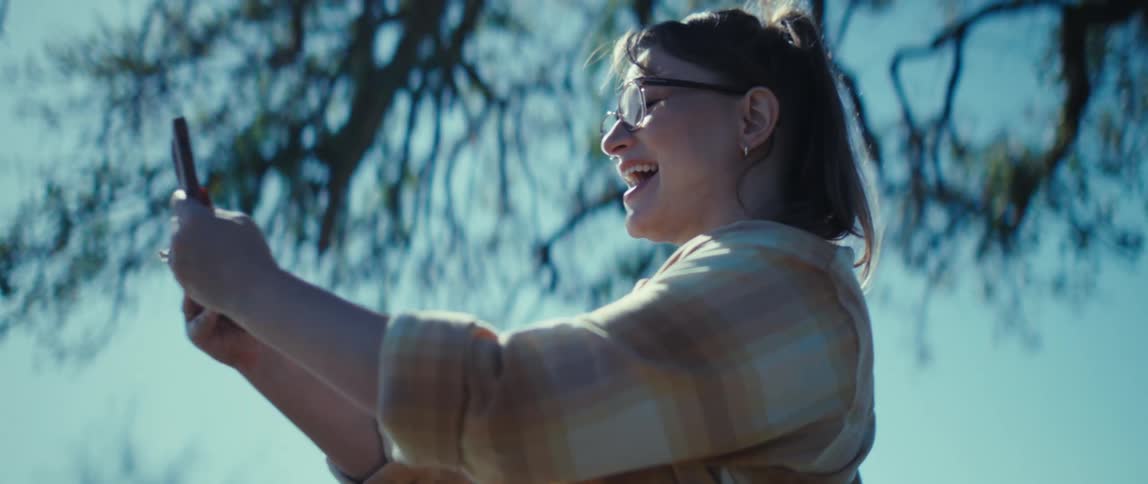 The woman is smiling and waving at her phone under the tree