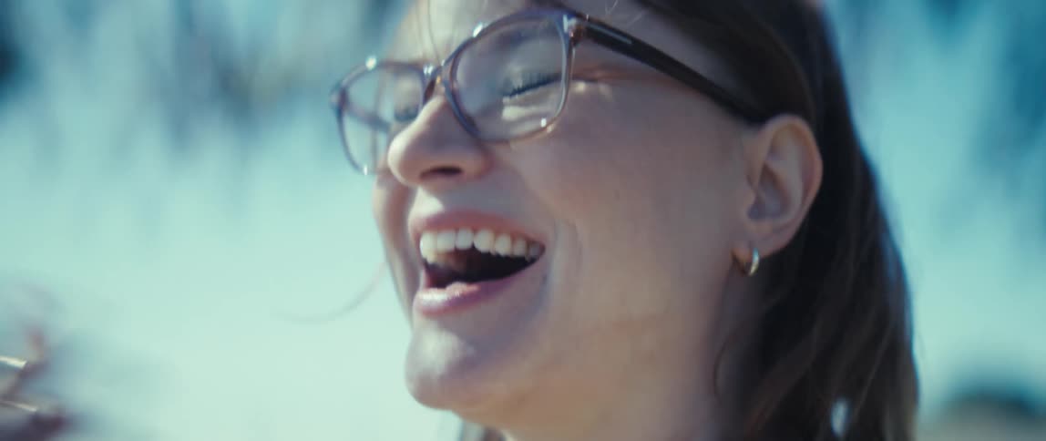Woman wearing glasses and light jacket holds phone with left hand against blue sky and blurred branches