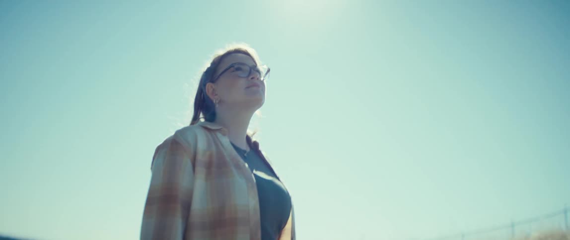 A woman wearing a plaid overshirt and eyeglasses stands on a grassy hill overlooking a body of water looking up at the bright blue sky with sun flare