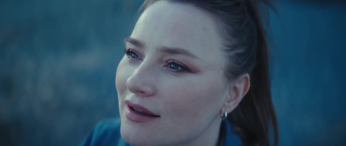 A womans face is seen in a closeup she looks toward the upper left corner of the frame her mouth opens slightly in a relief expression and a solid blue background fills the space behind her