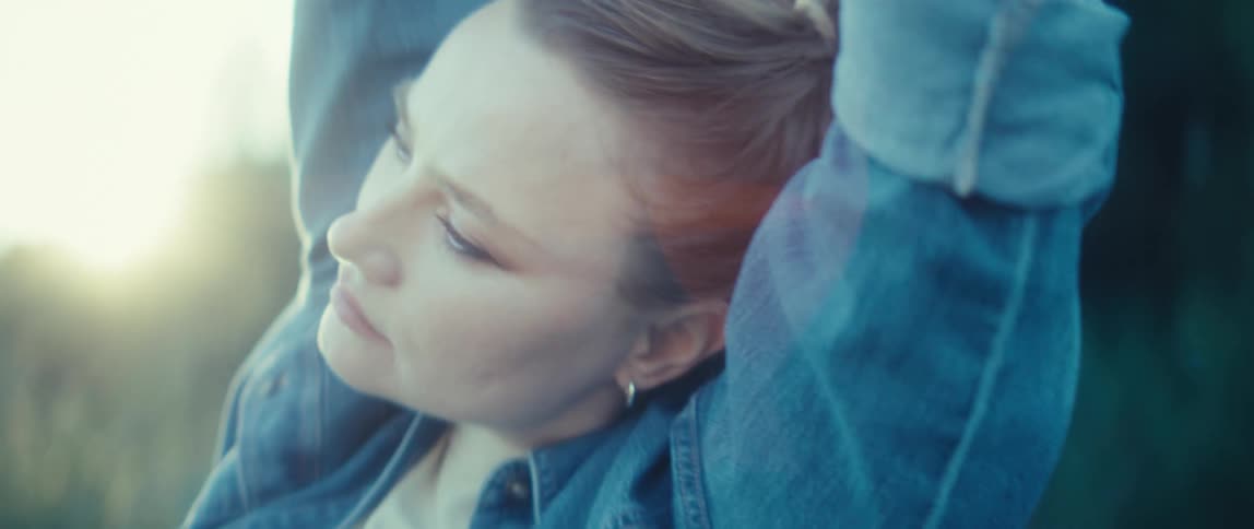 A woman wearing a denim jacket stands in profile she stretches her arms upward behind her head her face is tilted up slightly toward the bright sunlight and she has a calm expression