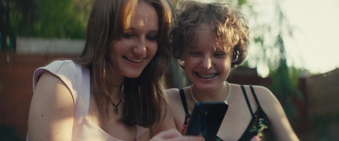 Two women sitting side by side look at phone screen in outdoor setting with wooden fence and greenery in the background