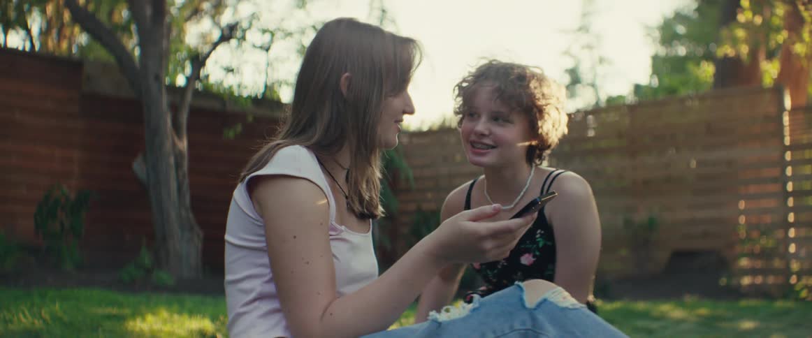 Two teenage girls sit on a lawn in a backyard scrolling through a mobile device and smiling at sunset