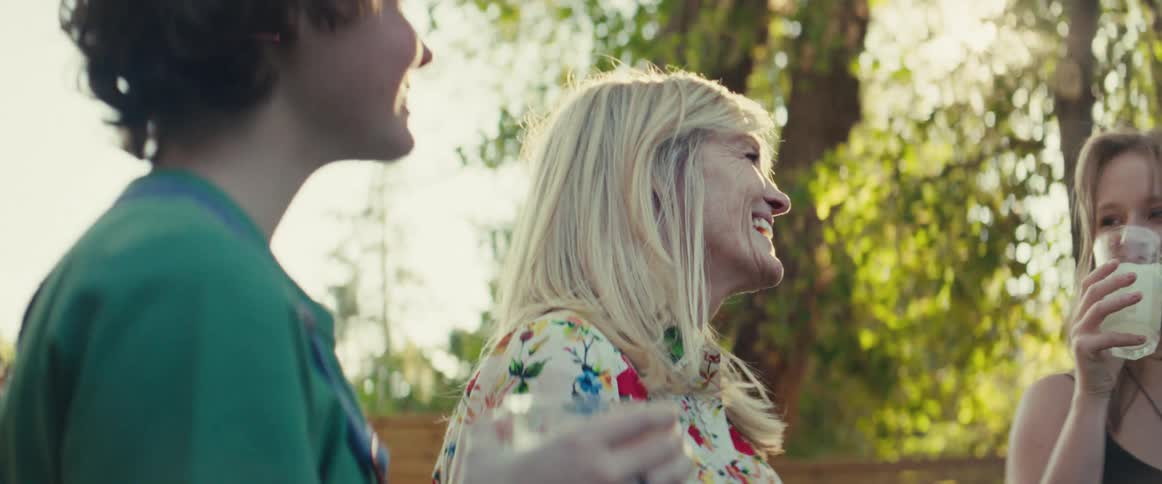 The video features a woman with long blonde hair wearing a white shirt with a colorful floral pattern laughing heartily in an outdoor sunny settingshe is surrounded by friends one of whom is holding a drink suggesting a joyful casual gather