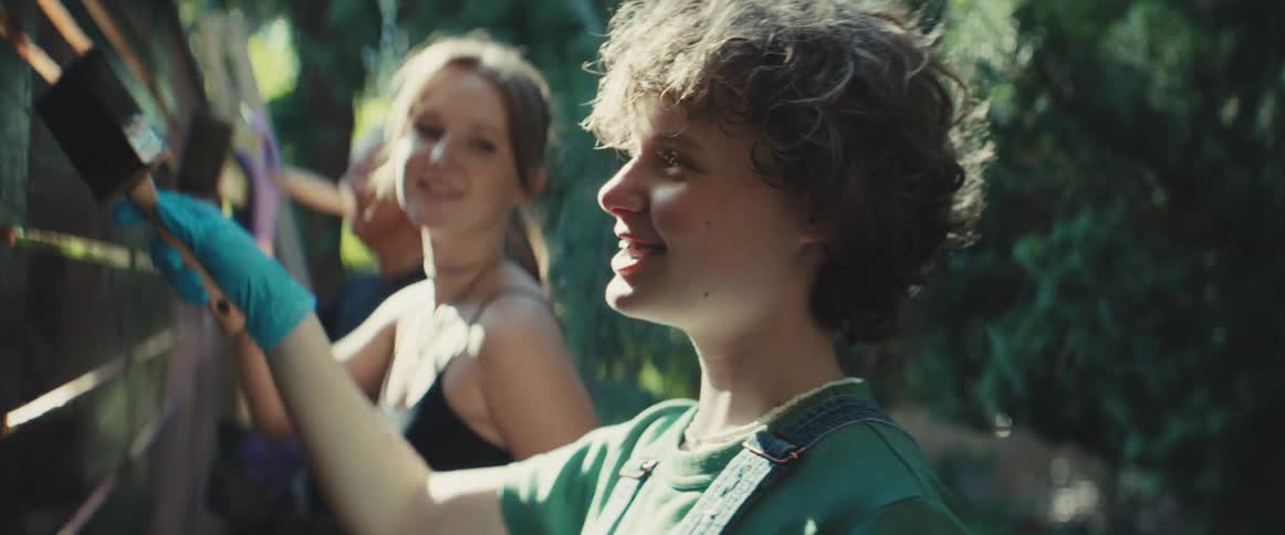 Teenagers wearing light clothing smiles and reaches upward with a gloved hand painting a wooden gate while another person smiles in the background and they are painting outdoors under sunlight