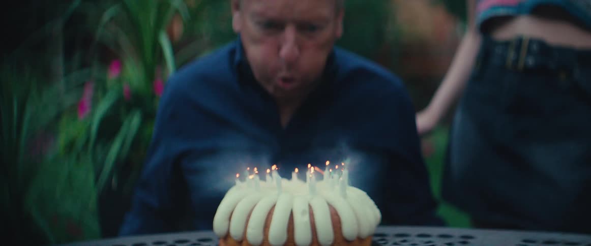 A man sits behind a table on the backyard lawn blowing out candles on a cake while celebrating his birthday at midday
