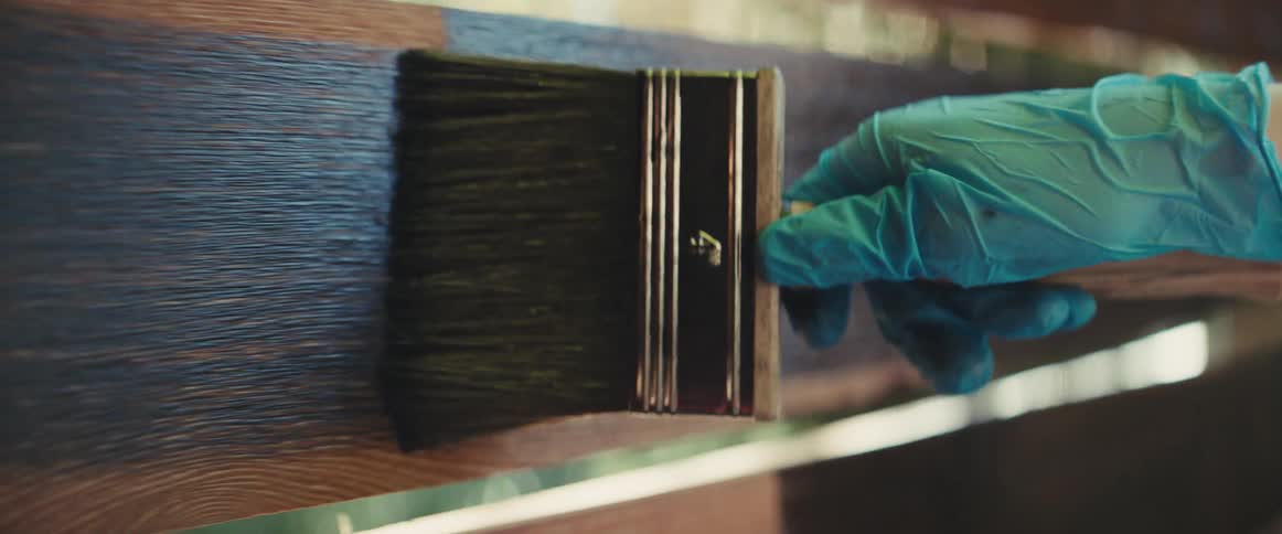 Person wearing a glove applies blue paint to a wooden board during the daytime