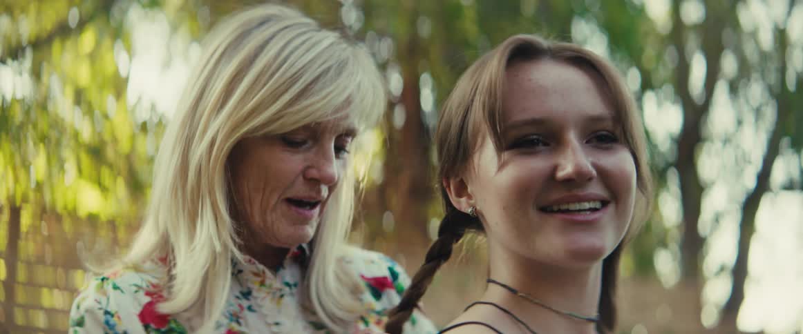 A woman braids a smiling girls hair outdoors on a sunny day