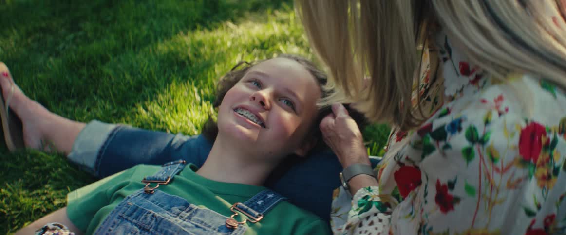 A smiling teen girl with braces lies on the grass in denim overalls looking up at an older woman who gently touches her hair