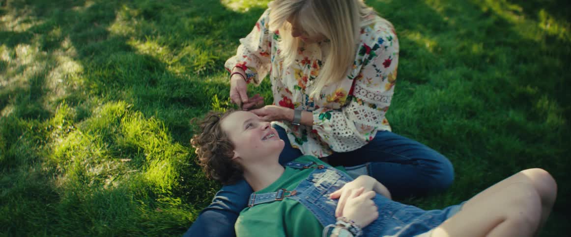 A person with blonde hair sits on green grass and touches the hair of another person lying with their head in the lap the person lying down looks up and smiles and the scene is set in bright sunlight