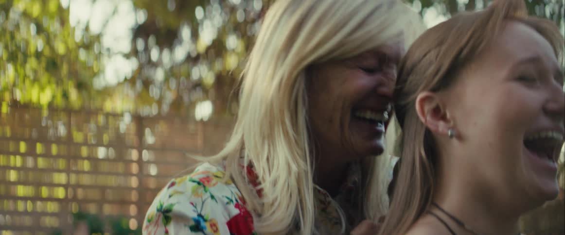 The video is a closeup of an older woman with long blonde hair smiling warmly outdoors in natural sunlight she is wearing a white shirt with a bright floral pattern and is interacting with a younger person whose back is to the camera