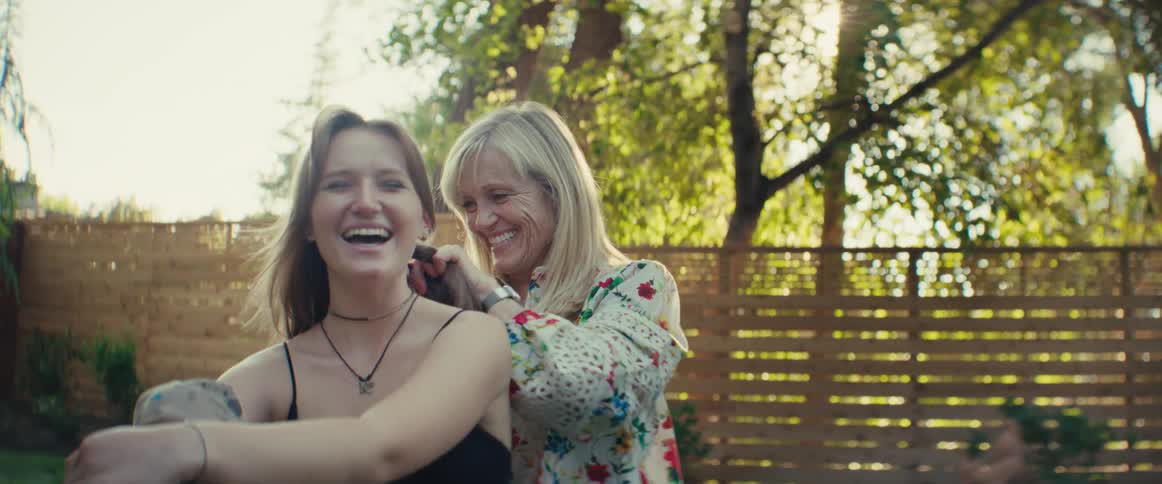 A laughing blonde woman in a white floral blouse gently adjusts the hair of a young woman sitting in front of her outside on a bright sunny day with a wooden fence and green trees in the background